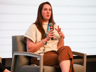 A woman with long brown hair, wearing a cream colored shirt and brown paints sits on a chair on a stage, holding a microphone, talking and gesturing with her hand.