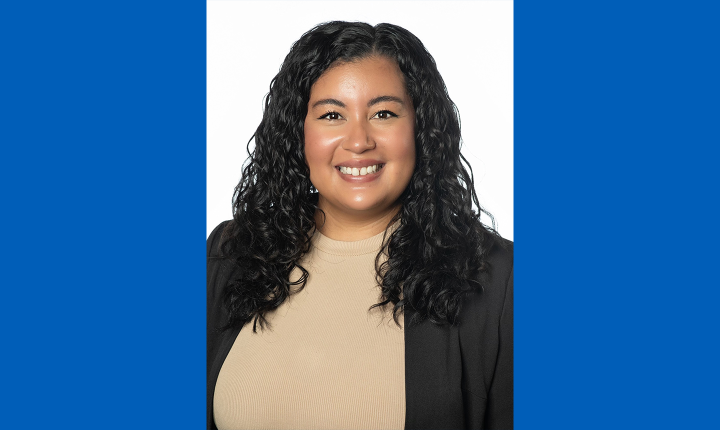 A portrait of Selina Tucker on a blue background. She has long black curly hair and is wearing a tan shirt with a black blazer over it.