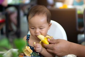 Toddler eating banana being fed by adult