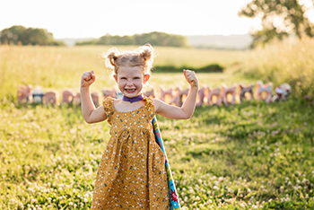 Quinn Liberman in a yellow sundress with a cape around her neck holding both her arms up to show her "muscles"