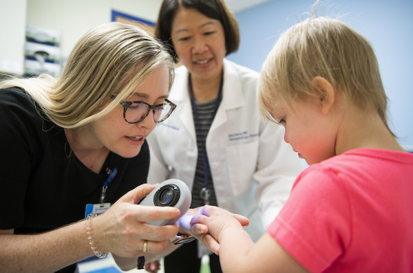 A young girl's hand is examined by a physician using a medical device while another physician observes.